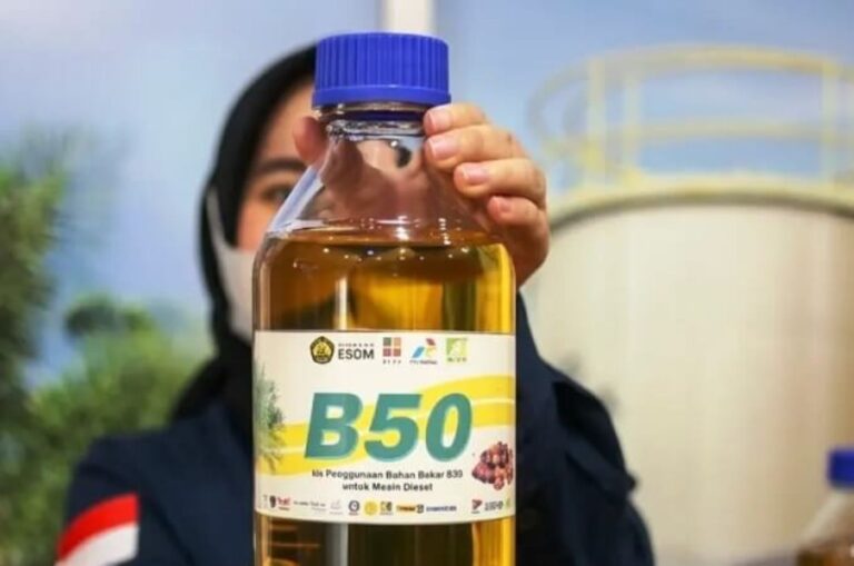 Person wearing a hijab holds up a large bottle with a blue cap labeled B50 biodiesel, in a workshop setting.