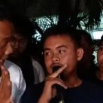 Three young men at night the center man holds a finger to his lips in a shushing gesture while the others look on