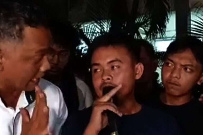Three young men at night; the center man holds a finger to his lips in a shushing gesture while the others look on.