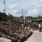 Rubble and collapsed roof tiles line a street after a disaster with a man on a bicycle passing by