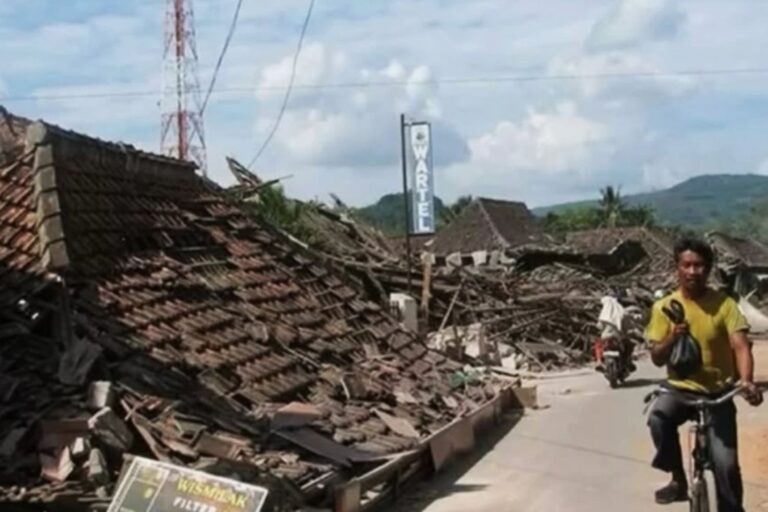 Rubble and collapsed roof tiles line a street after a disaster, with a man on a bicycle passing by.