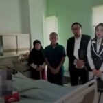 Four people stand beside a hospital bed in a clinical room with medical equipment nearby likely visiting a patient