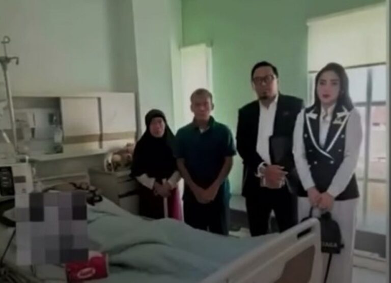 Four people stand beside a hospital bed in a clinical room with medical equipment nearby, likely visiting a patient.
