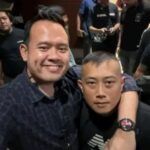Two men pose for a selfie in a crowded indoor setting one with his arm around the other and smiling at the camera
