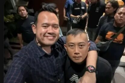 Two men pose for a selfie in a crowded indoor setting, one with his arm around the other and smiling at the camera.