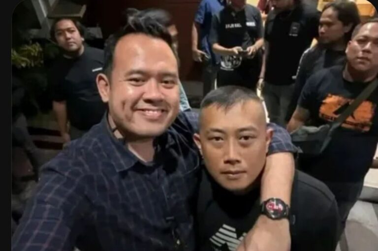 Two men pose for a selfie in a crowded indoor setting, one with his arm around the other and smiling at the camera.