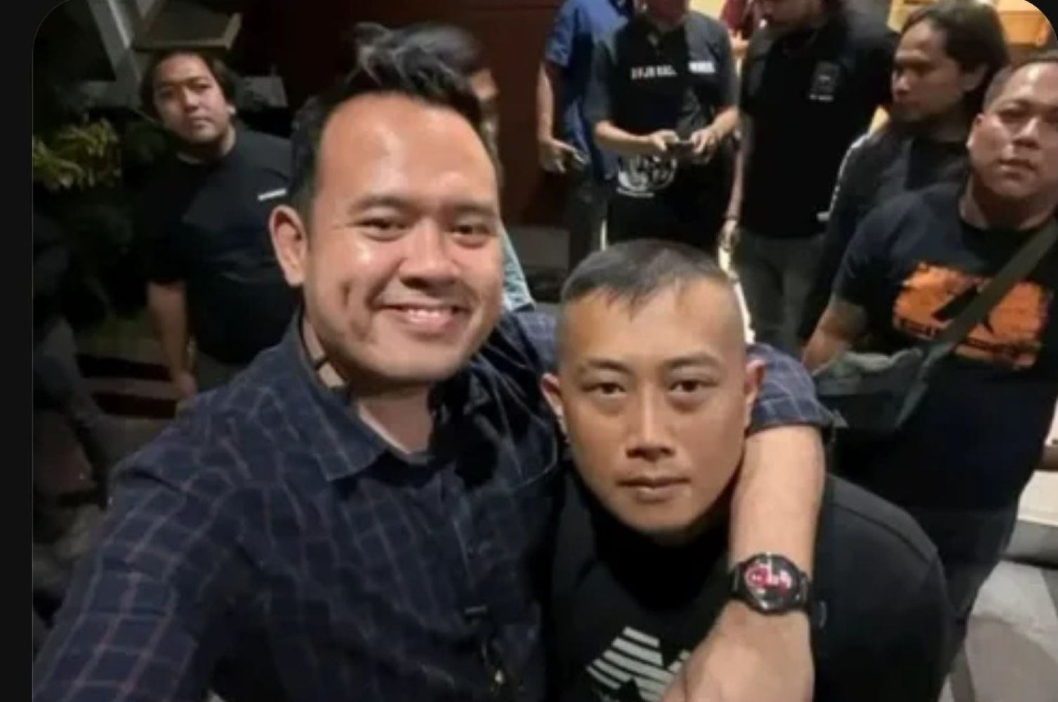 Two men pose for a selfie in a crowded indoor setting one with his arm around the other and smiling at the camera