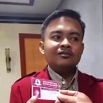 Man in a red blazer holding a pink business card toward the camera in an indoor setting facing slightly to the right
