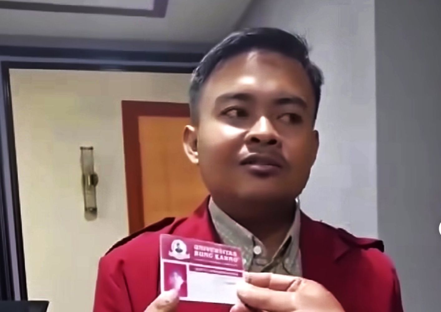 Man in a red blazer holding a pink business card toward the camera in an indoor setting facing slightly to the right