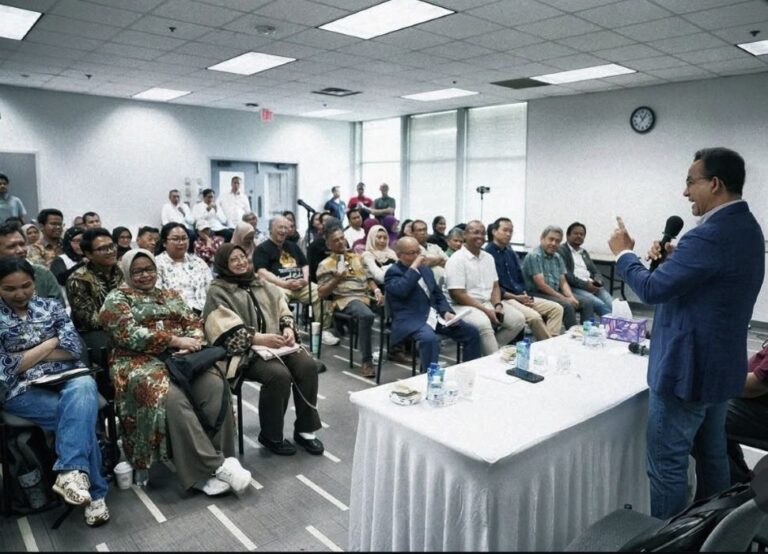 A speaker in a blue blazer addresses a diverse audience seated in a conference room, with a table of water bottles and microphones in front.
