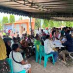 Crowd waiting under a canopy for a community aid event volunteers at a table assisting attendees