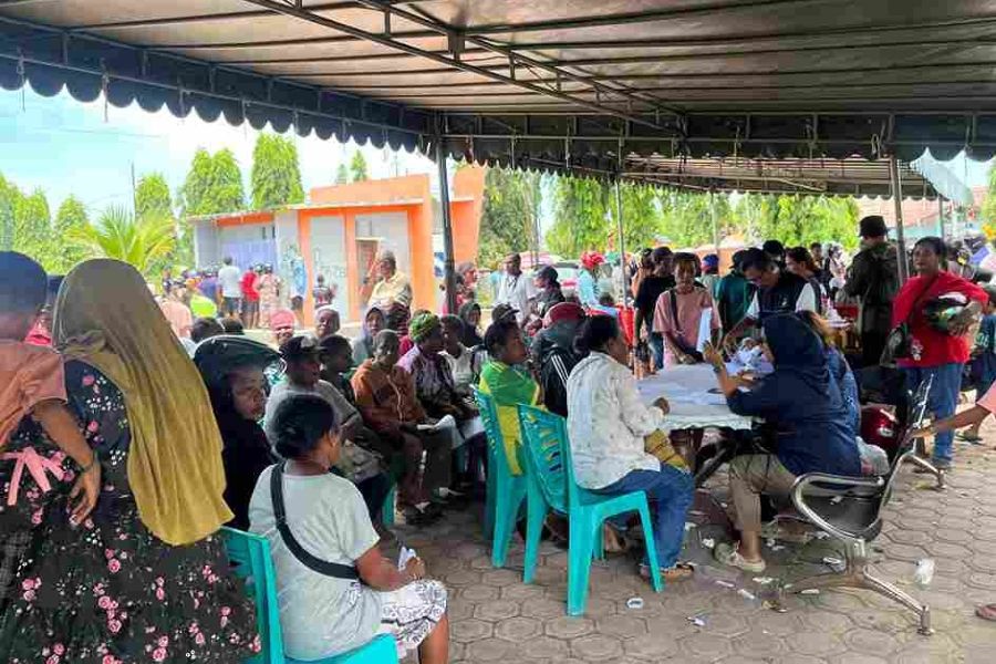 Crowd waiting under a canopy for a community aid event volunteers at a table assisting attendees
