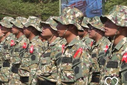 Line of soldiers in camouflage uniforms standing at attention with red ribbons on their chests under wide-brim hats.