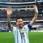 Soccer player in argentina jersey raising arms to celebrate on a packed stadium field after a match smiling at fans