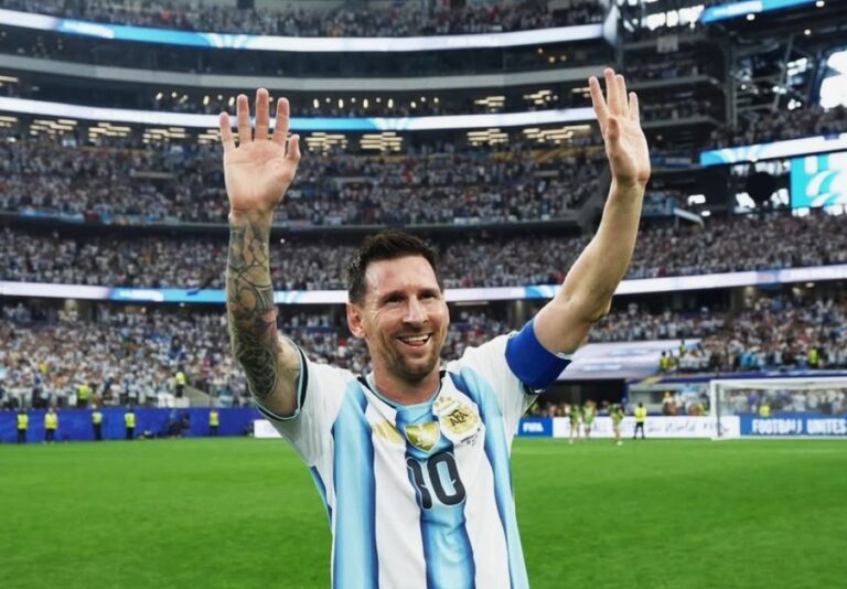 Soccer player in Argentina jersey raising arms to celebrate on a packed stadium field after a match, smiling at fans.