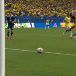 Soccer scene yellow clad players near the goal as a ball rolls toward the net goalkeeper in dark kit watches from the left stands filled with yellow clad fans