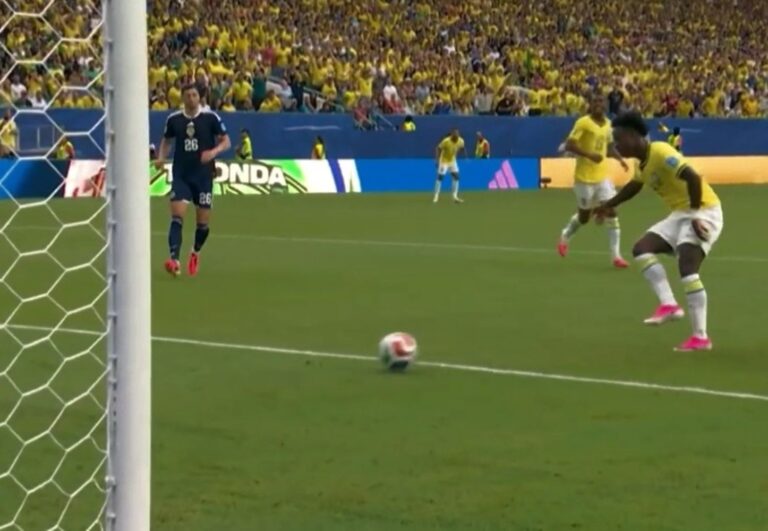 Soccer scene: yellow-clad players near the goal as a ball rolls toward the net; goalkeeper in dark kit watches from the left, stands filled with yellow-clad fans.