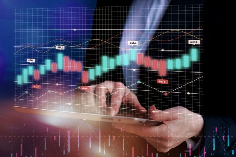Hands hold a tablet with a glowing stock chart overlay showing greenred candlesticks and buysell markers