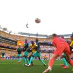 Soccer match near a goal as players in teal and yellow chase a ball in midair in a packed stadium