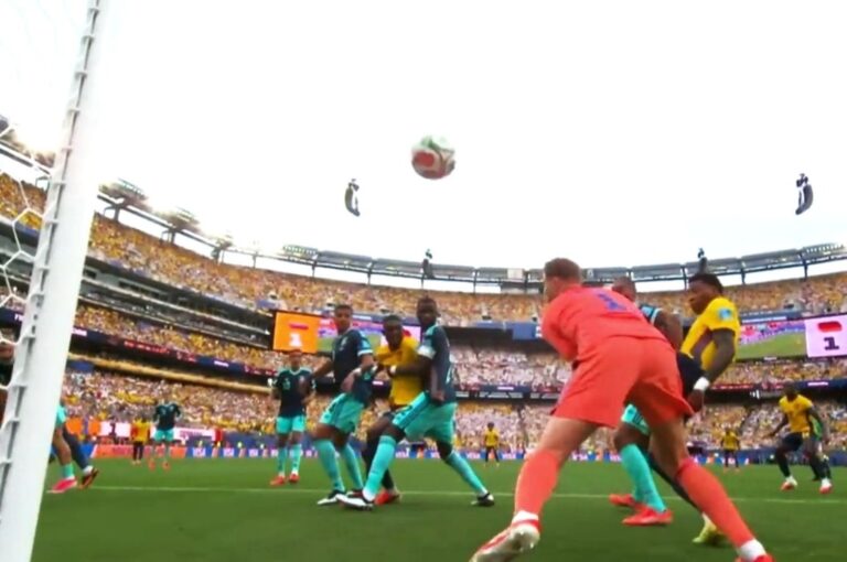 Soccer match near a goal as players in teal and yellow chase a ball in midair in a packed stadium.