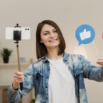 Smiling woman holds a selfie stick with a phone posing as social media like and heart icons appear beside her
