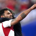 Two athletes hug in celebration on the field one in a white and red jersey raising his tattooed forearm and pointing upward in triumph