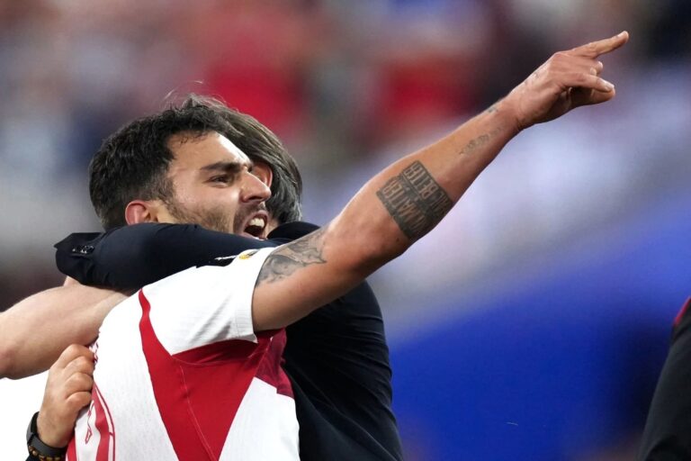 Two athletes hug in celebration on the field, one in a white and red jersey raising his tattooed forearm and pointing upward in triumph.