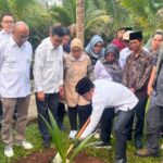 Group of people at a tree planting ceremony as a man in white bends to plant a sapling while others watch in a tropical garden