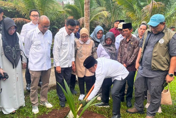 Group of people at a tree-planting ceremony, as a man in white bends to plant a sapling while others watch in a tropical garden.