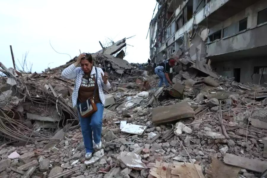 People walk through rubble of a collapsed building stepping over concrete and debris in a disaster scene