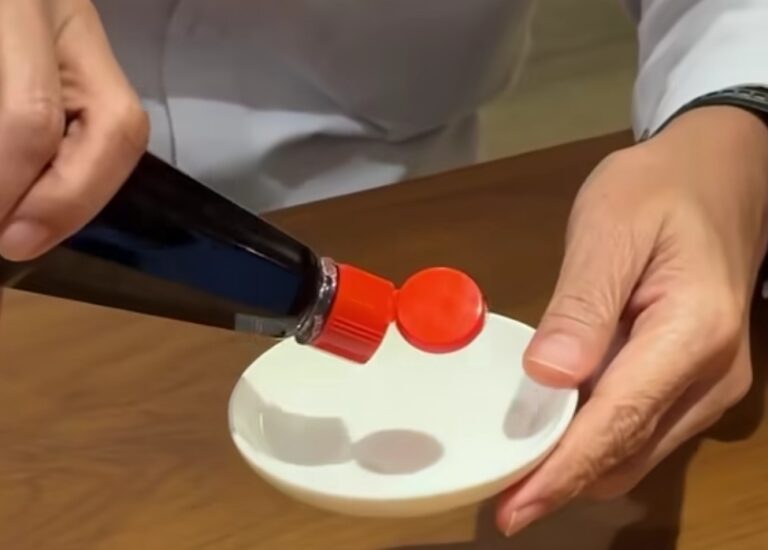 Person pours red liquid from a dark bottle onto a small white dish with a red cap at the edge of the plate.