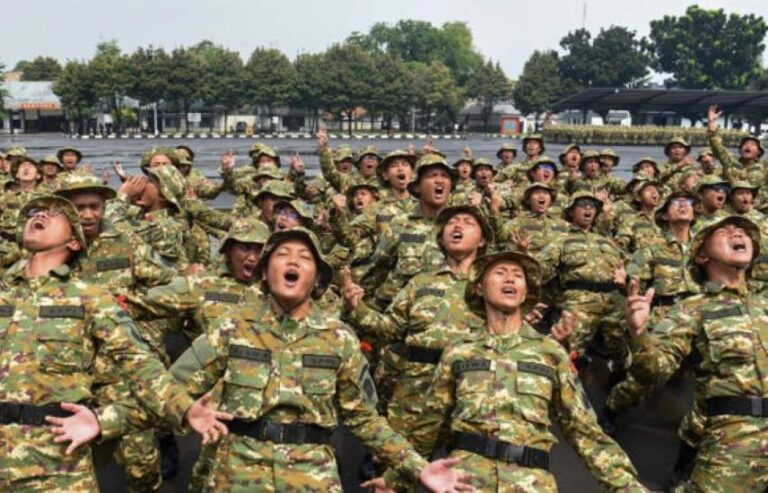 Large group of soldiers in camouflage uniforms outdoors, many mouths open and arms raised as if cheering or shouting during a drill.