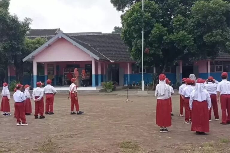 Group of students in red pants and white shirts stand in lines on a schoolyard, facing a central instructor as a microphone stand sits ahead; pink school building in background.
