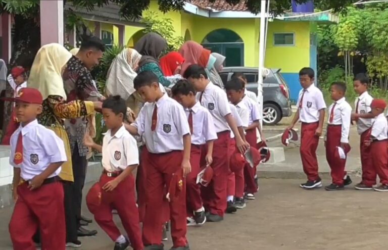 Group of schoolchildren in white shirts and red pants lined up outdoors, hands extended to receive items from adults wearing colorful headscarves and coats at a school event.