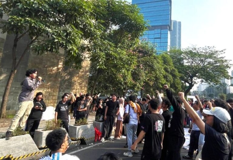 A street protest with a man speaking into a microphone on a raised platform as a crowd raises fists nearby on a sunny day in a city setting.