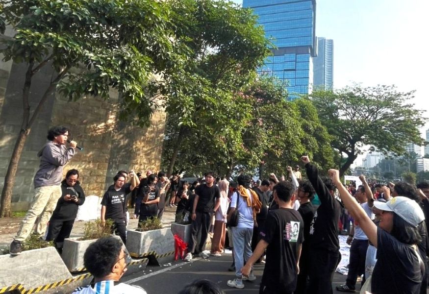 A street protest with a man speaking into a microphone on a raised platform as a crowd raises fists nearby on a sunny day in a city setting