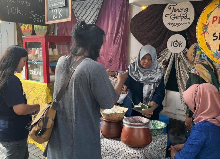 Women at a street market stall serving bowls of food, price signs show 10K in the background, warm daytime scene.