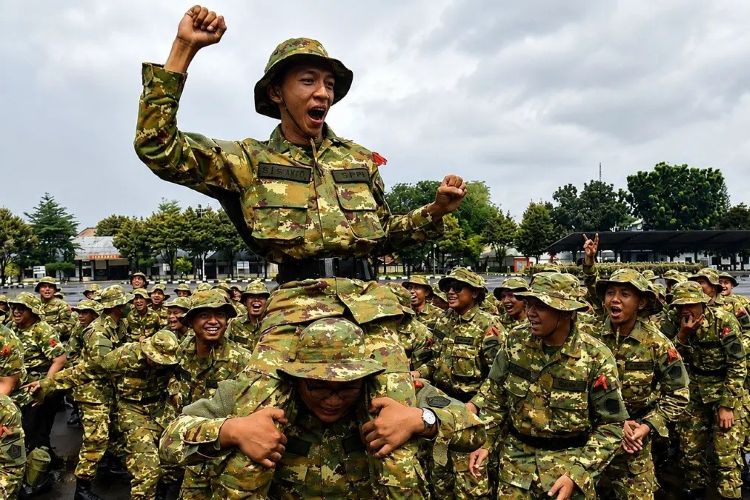 Soldier raised on teammates shoulders cheering with a fist in the air among a group of camouflaged soldiers outdoors