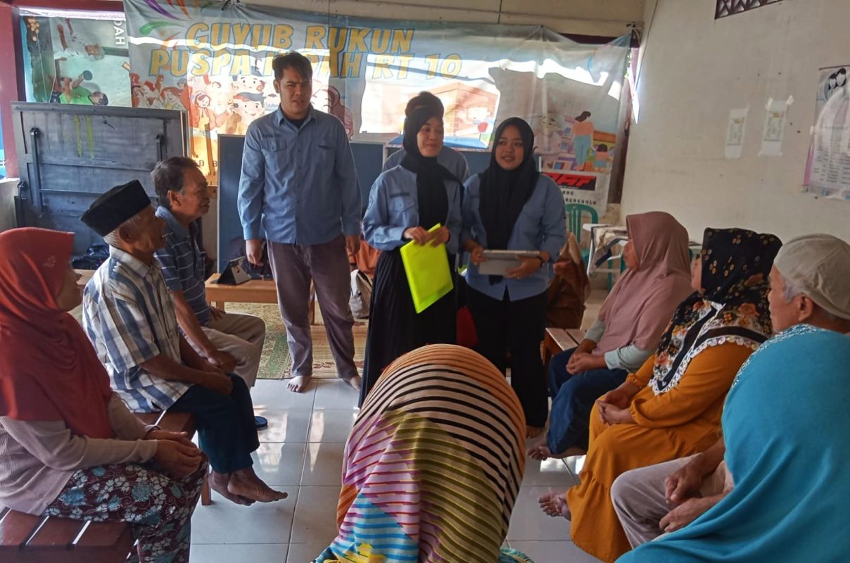 Group of villagers seated in a circle as two young women and a man in blue shirts present information at the front of a room Behind them a colorful banner hangs on the wall