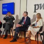 Panel of four professionals seated on stage in a discussion pwc logo visible behind them and a blue screen reading navigating global shifts on the left