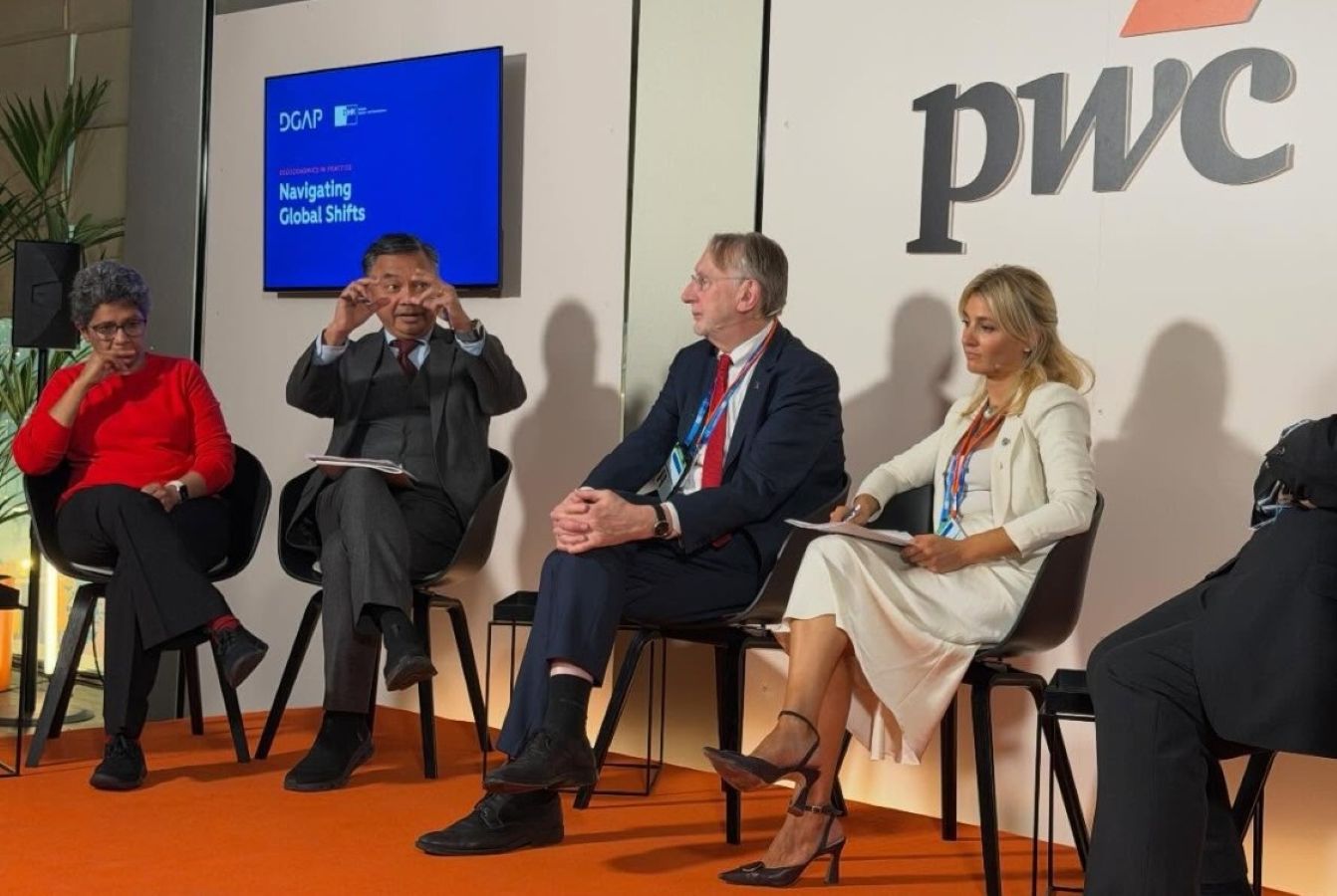 Panel of four professionals seated on stage in a discussion pwc logo visible behind them and a blue screen reading navigating global shifts on the left