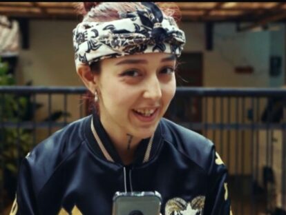 Young woman wearing a patterned headscarf and dark jacket smiles at the camera holding a phone