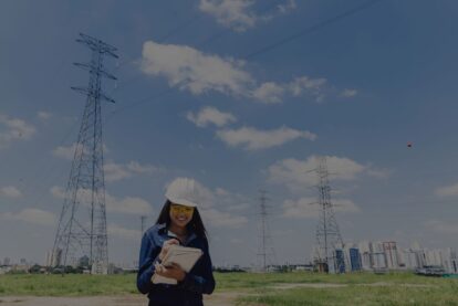 Woman in a white hard hat and yellow safety glasses writing in a notebook at a rural power transmission site with tall lattice towers and a distant city skyline