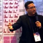 Male presenter in a black blazer speaks into a microphone in front of research posters with red bar charts behind him