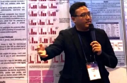 Male presenter in a black blazer speaks into a microphone in front of research posters with red bar charts behind him