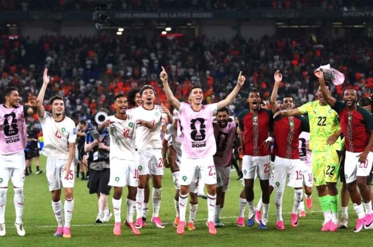 Soccer team celebrates on the field after a match, players in white kits with pink bibs and a neon‑yellow goalkeeper, crowd in the stands behind them.