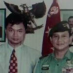 Two men posing for a photo one in a light shirt and patterned tie the other in a green military uniform with medals with portraits and a flag in the background