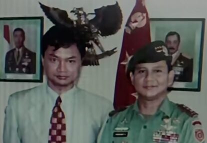 Two men posing for a photo one in a light shirt and patterned tie the other in a green military uniform with medals with portraits and a flag in the background