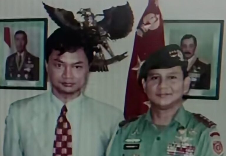 Two men posing for a photo: one in a light shirt and patterned tie, the other in a green military uniform with medals, with portraits and a flag in the background.
