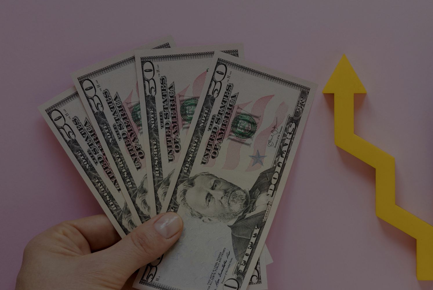Fanned stack of $50 bills held in a hand with a yellow upward arrow symbolizing financial growth